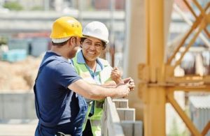 Two construction workers wearing safety gear and hard hats talking on site near scaffolding and equipment.