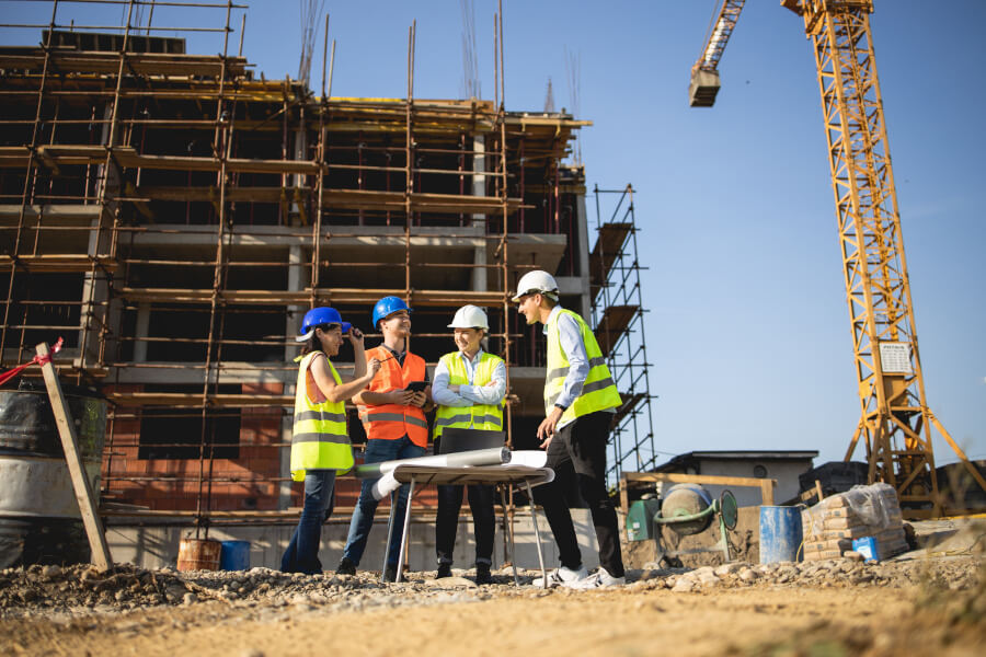 Group of construction professionals reviewing blueprints on-site in front of a scaffolded building under construction, with a crane in the background.