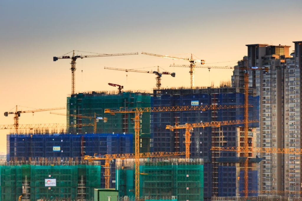 High-rise construction site with multiple tower cranes and partially completed buildings covered in scaffolding and netting at sunset.